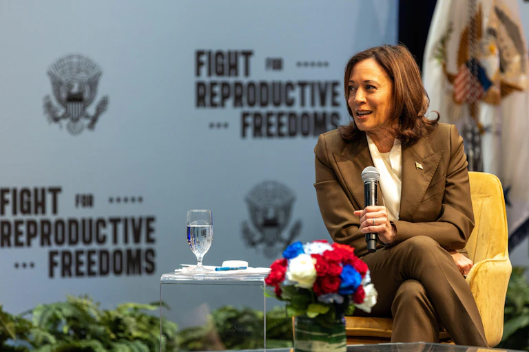 Kamala Harris sits speaking into a microphone in front of a backdrop with messages reading: FIght for Reproductive Freedom.