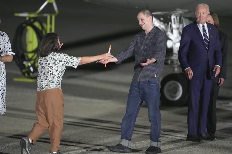 A man walks toward a woman to embrace her. A man in a dark blue suit stands behind them and smiles.