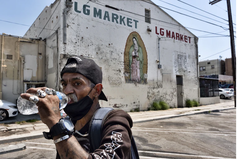 A man drinks water from a bottle while walking through a neighborhood with no trees in sight and lots of concrete and pavement.