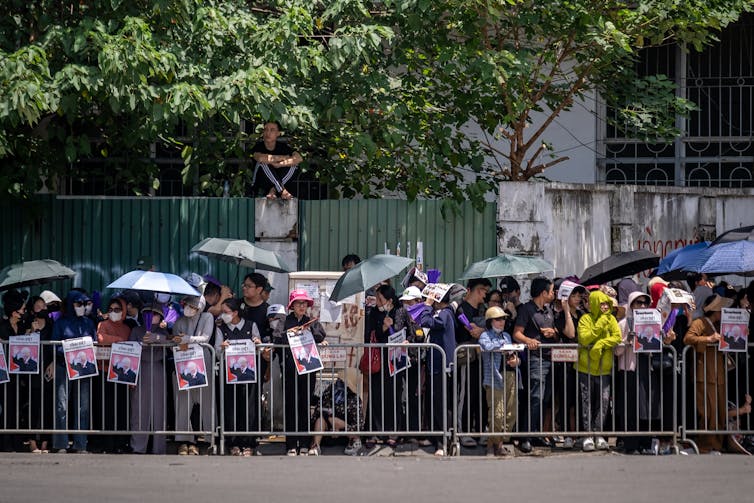 People line up behind metal barriers, many holding umbrellas.