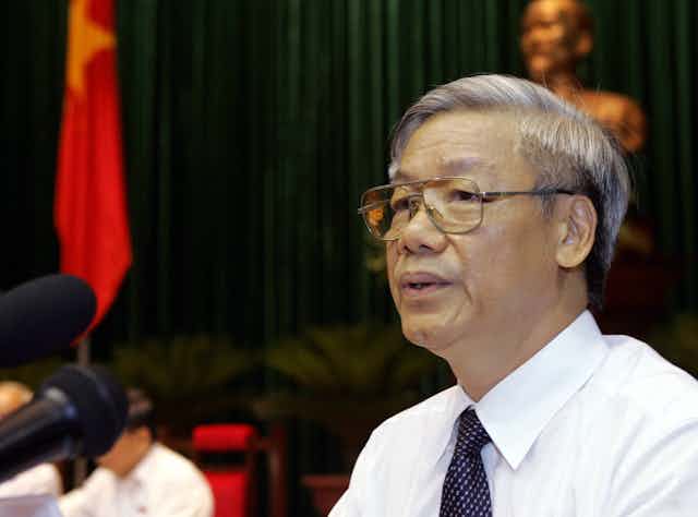 A man in glasses talks in front of a brass bust.