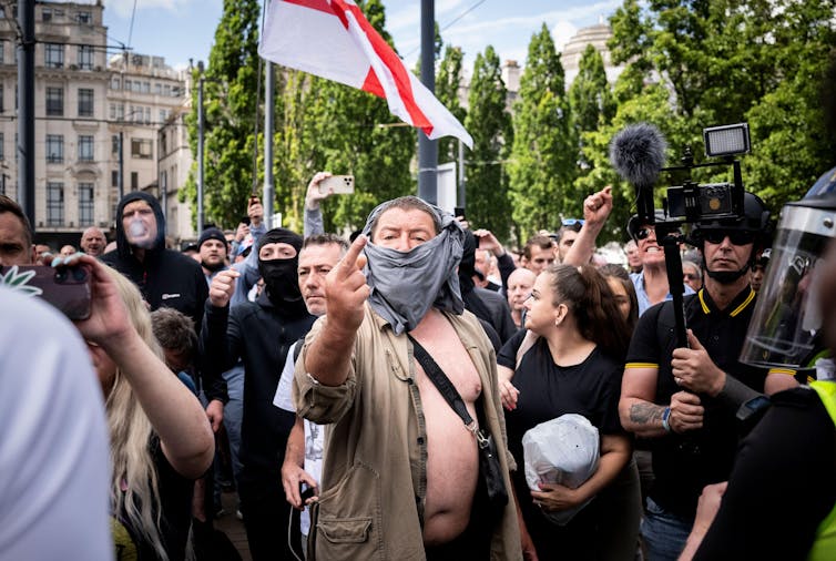 A man with a face covering standing in front of a crowd carrying an English flag gives the middle finger to the camera.