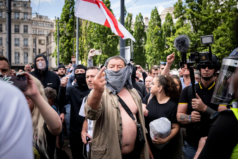 A man with a face covering standing in front of a crowd carrying an English flag gives the middle finger to the camera.
