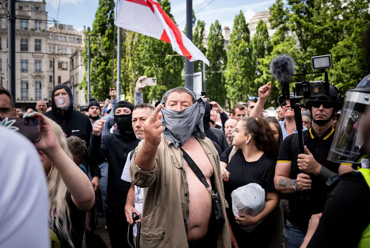 A man with a face covering standing in front of a crowd carrying an English flag gives the middle finger to the camera.