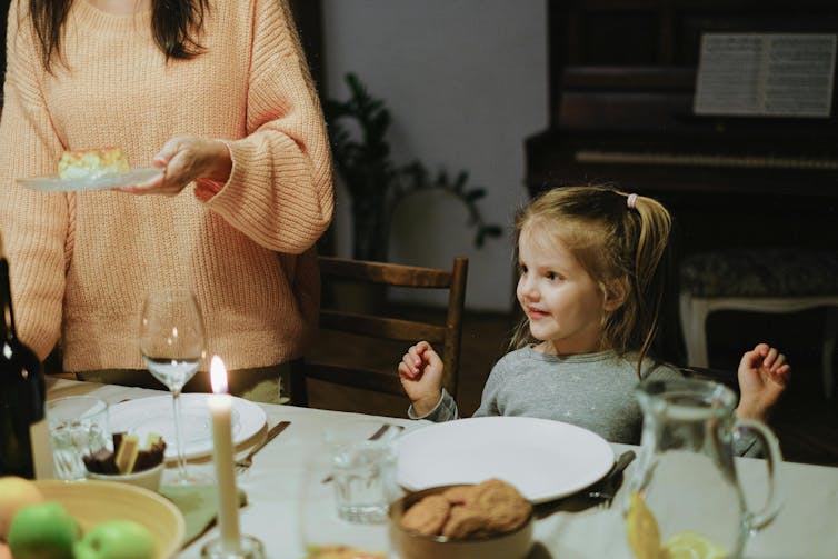 A young child sits at a dining table. She looks excited as an adult serves a plate of food.