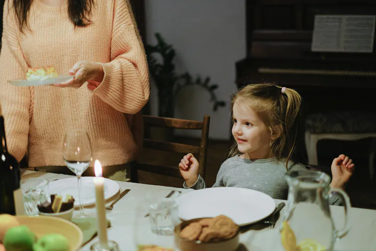 A young child sits at a dining table. She looks excited as an adult serves a plate of food.