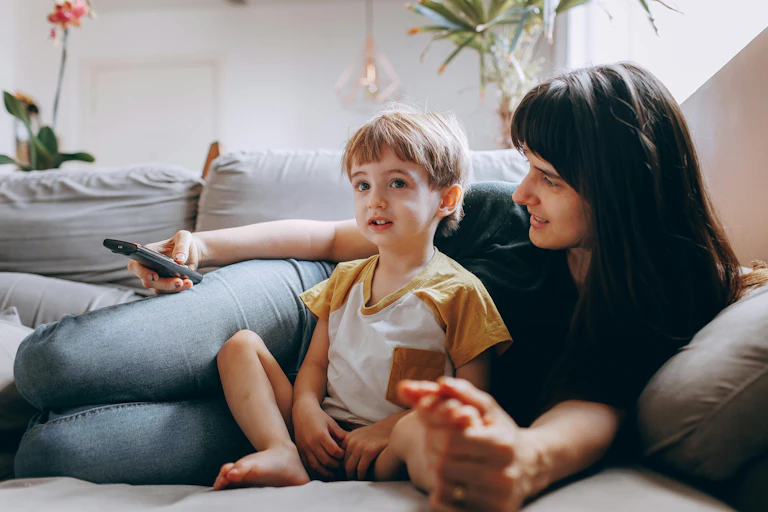 A woman lies on a couch with a TV remove. A young child is propped up in her lap.