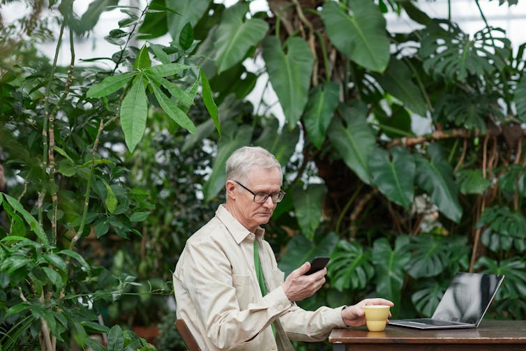 An older man sits alone but is looking at his phone while also using his laptop.