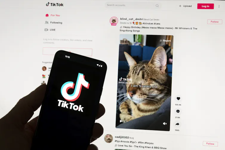 TikTok logo is seen on a mobile phone in front of a computer screen which displays the TikTok home screen.