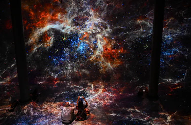 Two people photograph a vivid blue, orange and white image captured by a space telescope