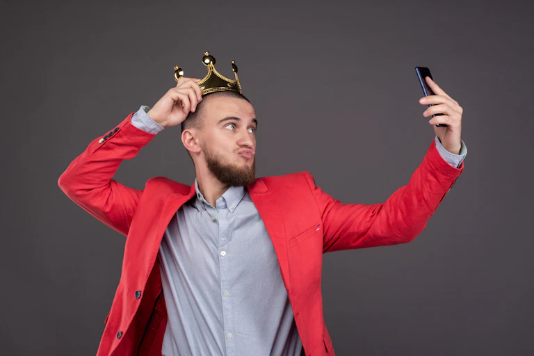 Young bearded handsome man in gold crown taking selfie looking at smartphone..