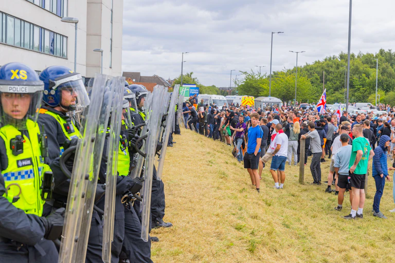 Side view of a row of police in riot gear protecting the hotel, while a crowd of protesters stands opposite them