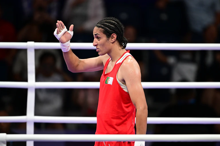 A woman in a red vest with cornrows in her hair stands in a boxing ring and raises a bandaged hand.