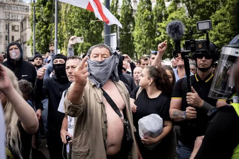 Un homme avec un foulard qui le masque fait un doigt d'honneur au milieur d'une foule composée de journalistes et d'autres hommes et femmes masqués