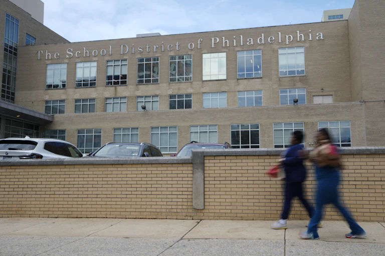 Two pedestrians walk past a large building with lettering that reads: The School District of Philadelphia