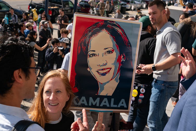 A white woman holds up a red, white and blue poster that has a drawn image of a woman's face with the word 'Kamala' below it.