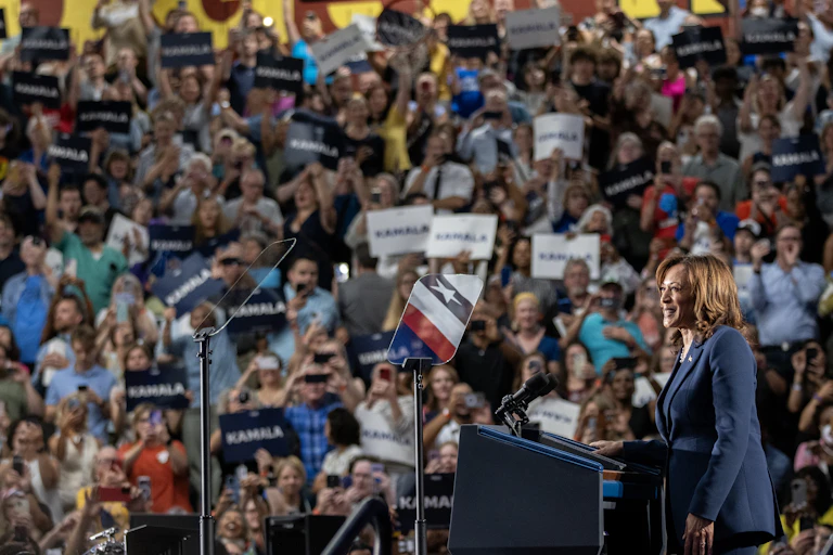 A woman in a blue pantsuit stands at a lectern in front of a large crowd of people, many of whom hold posters that say 'Kamala.'