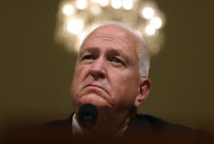 A stern-faced, middle-aged white man listens to a question during a congressional hearing.