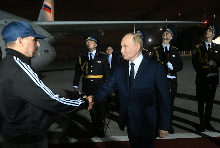 A man in a blue hat shakes hands with a man in a suit, an airplane can be seen in the background.