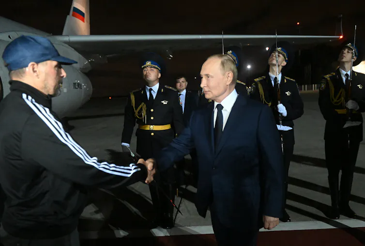 A man in a blue hat shakes hands with a man in a suit with an airplane in the background.
