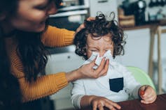An adult holds a tissue to a child's nose, helping them cover their sneeze