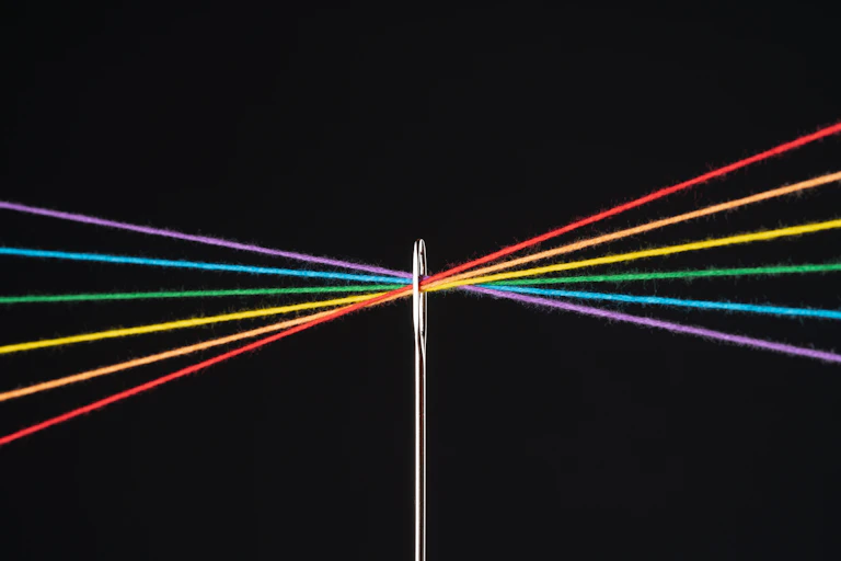A needle with threads the colors of the rainbow running the eye over a black background.