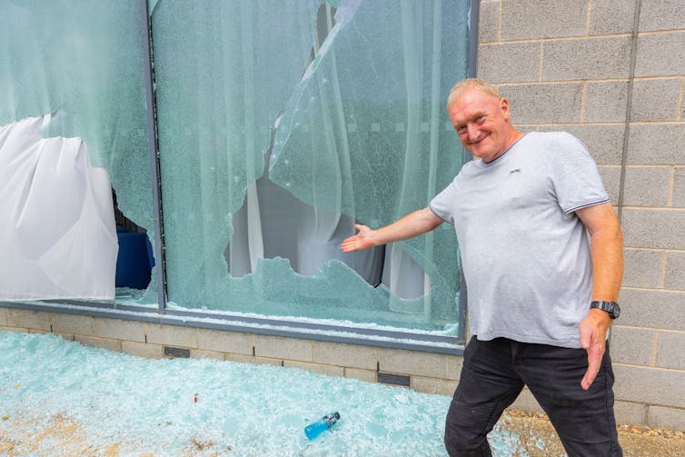 Um homem sorrindo em frente a uma janela quebrada, estendendo o braço como se quisesse mostrá-la como uma exposição.
