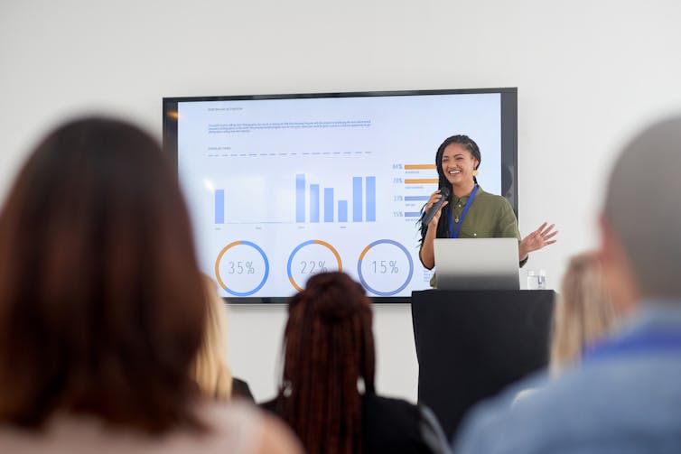 woman at podium with slides beside her, presenting to a room