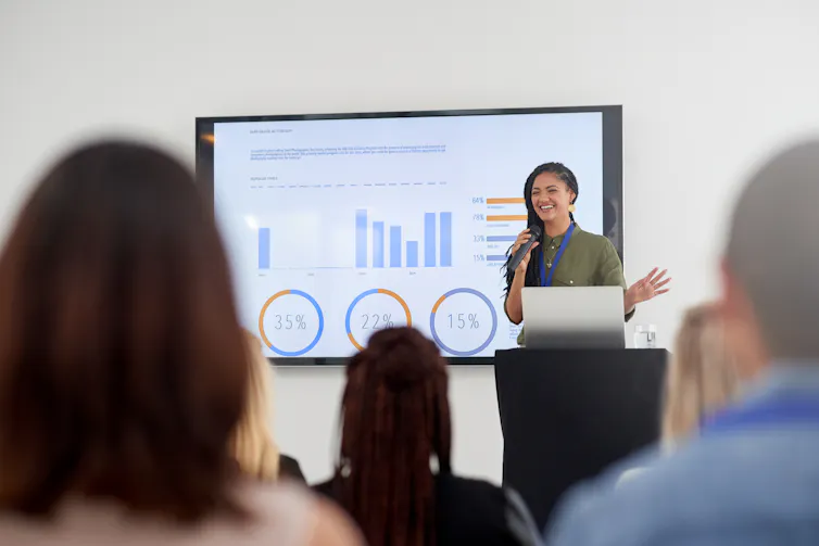 woman at podium with slides beside her, presenting to a room