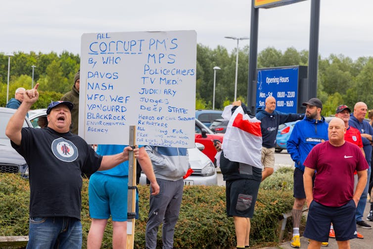 A man in a group holds up a sign referencing various conspiracy theories. Another wears an English flag over his shoulders.