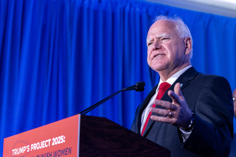 An older white man with white hair stands at a podium and wears a dark suit with a red tie.