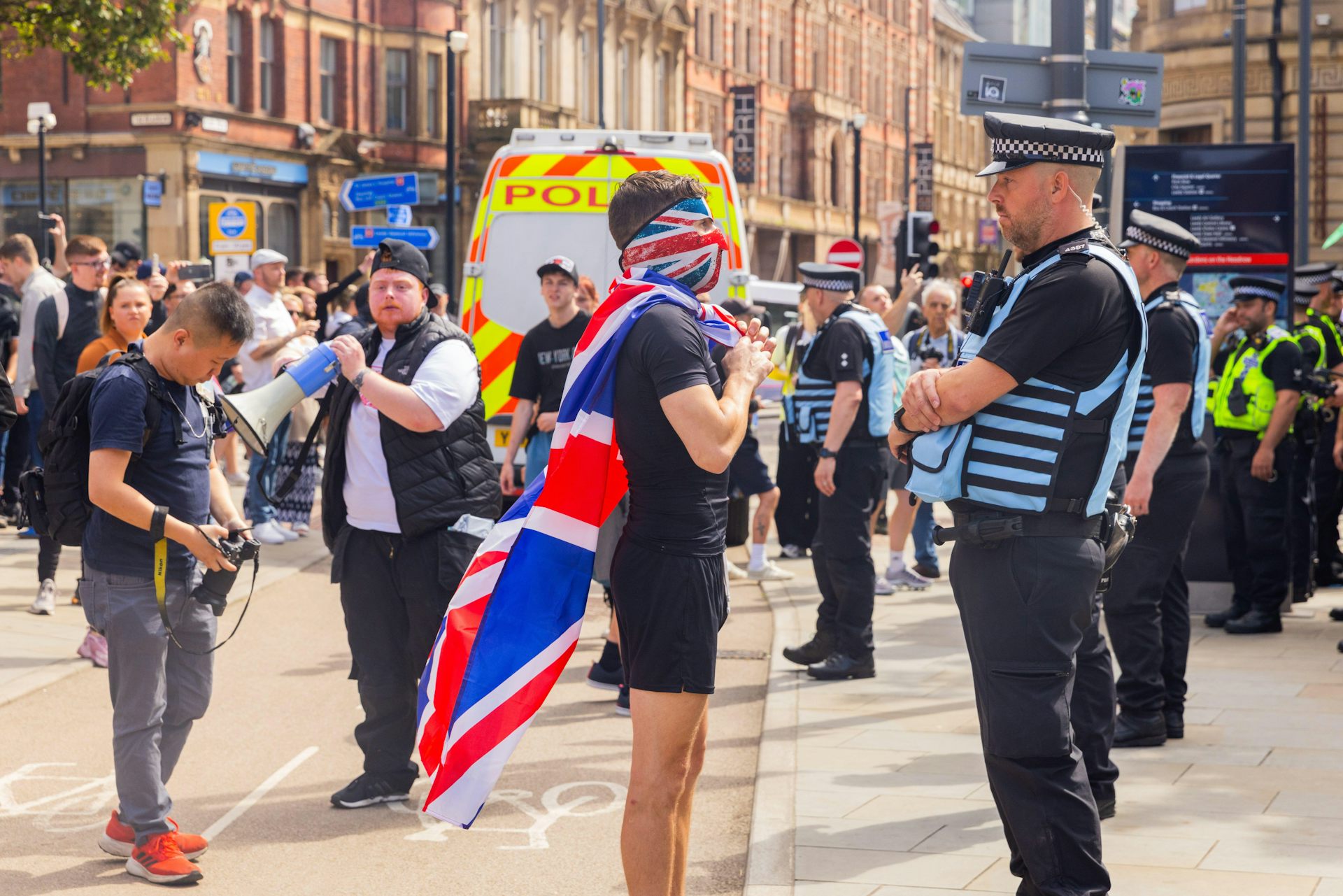 Um homem usando uma máscara com a bandeira do sindicato e outra bandeira do sindicato pendurada nas costas como uma capa fica frente a frente com um policial.