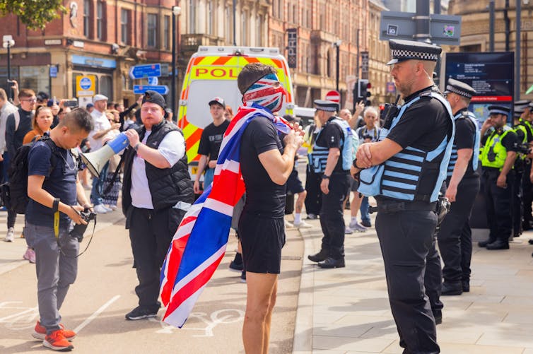 Um homem usando uma máscara com a bandeira do sindicato e outra bandeira do sindicato pendurada nas costas como uma capa fica frente a frente com um policial.