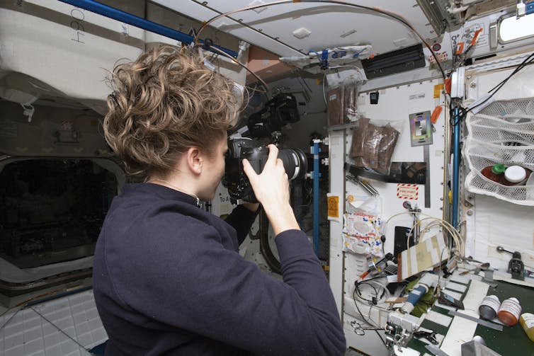 A woman is seen from behind in a module of the International Space Station. She holds a camera up to her face to take a picture of the wall. Her hair is loose and floating because of the lack of gravity.