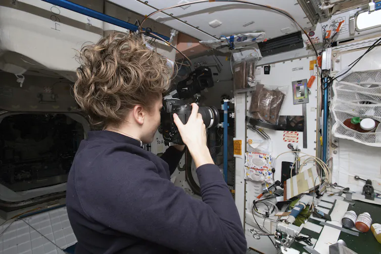 A woman is seen from behind in a module of the International Space Station. She holds a camera up to her face to take a picture of the wall. Her hair is loose and floating because of the lack of gravity.