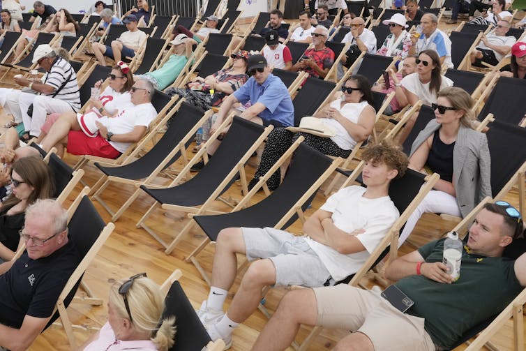People sit on deck chairs waiting for a tennis game amid empty seats.