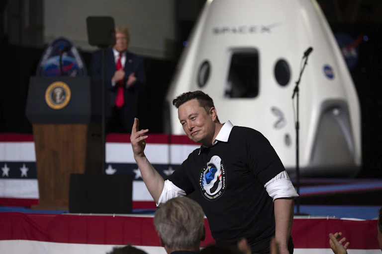 Elon Musk stands with his hand in the air while behind him on a stage, Donald Trump claps next to a SpaceX capsule.