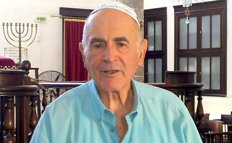 A bald man in a light-blue shirt and white skullcap stands near a menorah.