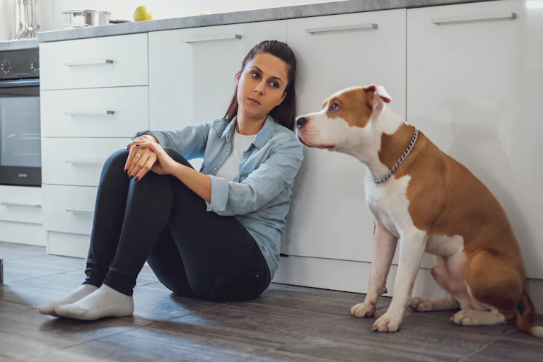 A sad looking woman sitting on a kitchen floor next to a dog.