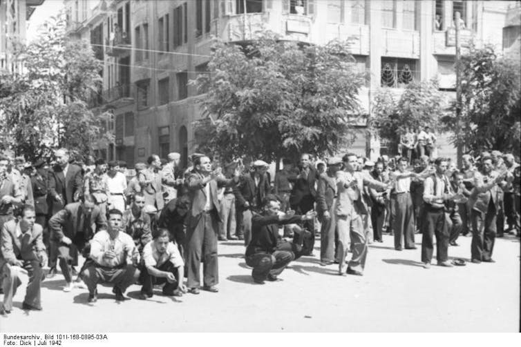 A black and white photograph of a crowd outdoors, with a few rows of men looking bewildered and doing exercises.