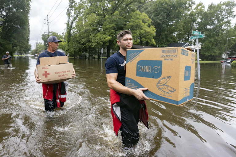Two firefighters wade through knee-deep water carrying large boxes. The streets and surrounding yards are flooded.