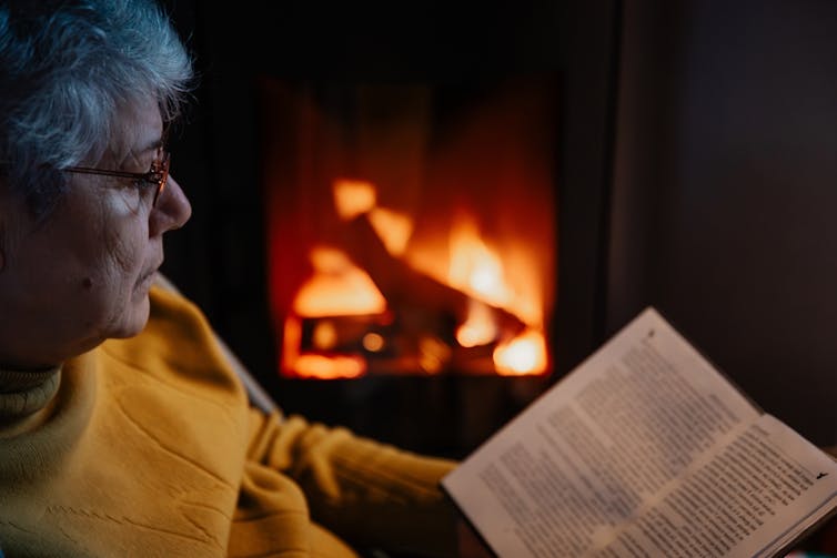 Older woman sitting reading book next to wood fire