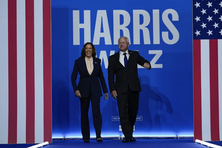 A dark-haired woman in a blue suit and a balding man smile as they walk on a stage. a banner with the words harris walz is behind them.