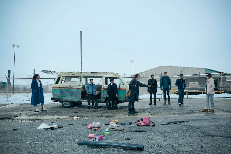 The Umbrella Academy character stand in an empty lot by a wrecked van.