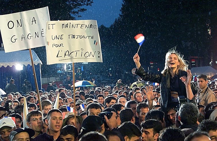 A crowd celebrates, with a smiling young blonde woman on someone's shoulders waving a small French flag.