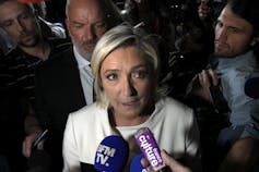 A blonde woman in a white suit jacket speaks into recording devices held in front of her by reporters.