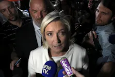 A blonde woman in a white suit jacket speaks into recording devices held in front of her by reporters.