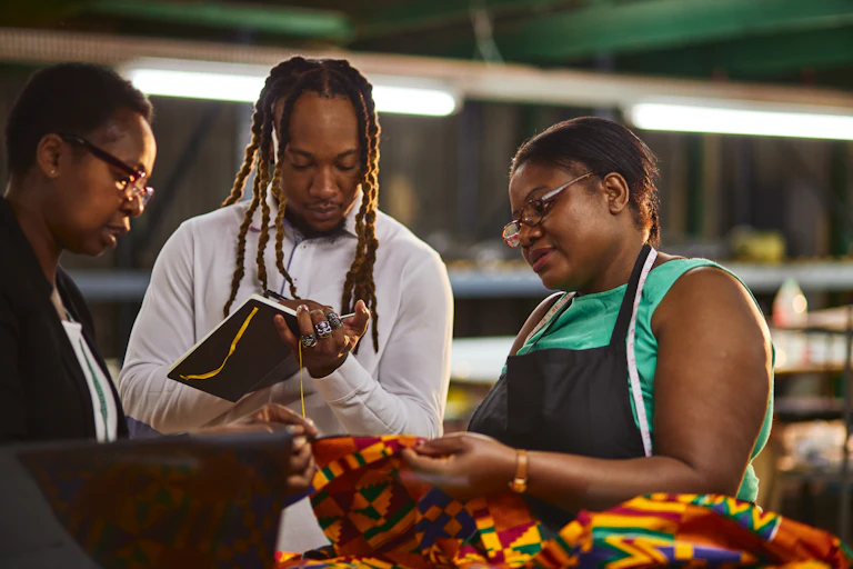 Two women look at a piece of fabric while a man makes notes in a book