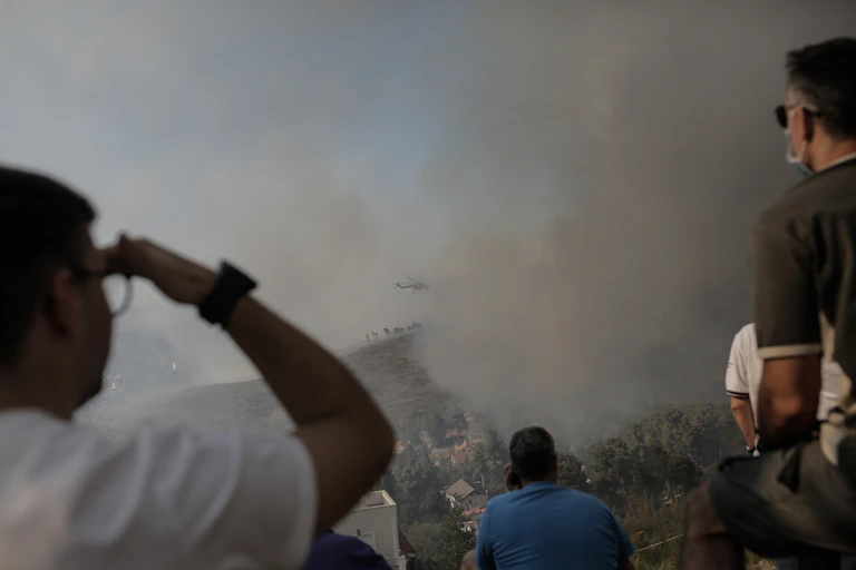 People watch a firefighting helicopter as smoke rises in the distance.