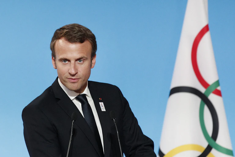 Man speaks with Olympic flag in background.